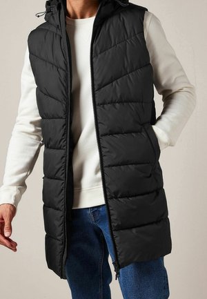 Mężczyzna nosi długą czarną pikowaną kamizelkę bez rękawów na białym swetrze i niebieskich dżinsach, z ręką w kieszeni i częściowo rozpiętym zamkiem błyskawicznym.