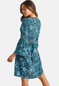 Robe turquoise avec motif floral bleu, manches longues avec poignets évasés, et corsage ajusté, présente une taille froncée et une texture lisse.