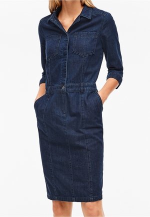 Femme portant une robe-chemise en denim bleu foncé avec des manches trois quarts, des poches avant et des poches latérales, debout, les mains dans les poches.