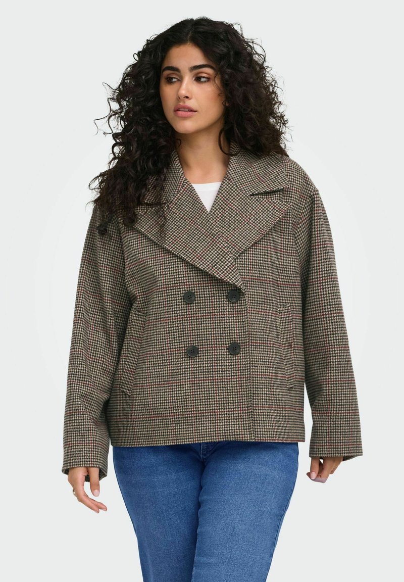 Femme aux cheveux bouclés porte une veste croisée à carreaux marron et un jean bleu, regardant légèrement vers sa gauche sur un fond uni.