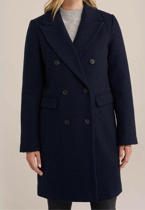 Femme portant un manteau en laine bleu marine foncé à double boutonnage avec des revers crantés, par-dessus un pull gris clair.