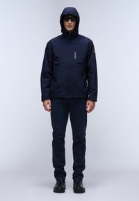 Marineblauwe weerbestendige jas met een capuchon, rits aan de voorkant, twee zijzakken en een logo-detail op de borst. Gecombineerd met bijpassende broek.