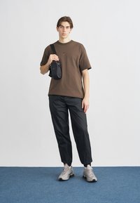 T-shirt di cotone marrone con piccolo logo, borsa tracolla in nylon nero, pantaloni neri a gamba stretta e sneakers grigio chiaro su un tappeto blu.