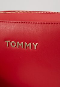 Czerwona skórzana torba na zamek błyskawiczny w kolorze złotym z metalowymi literami wypisującymi "TOMMY" na przedniej powierzchni.