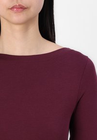 Burgundy långärmad topp med båtringning, slät textur och figurnära design. Tyget verkar mjukt och något elastiskt.