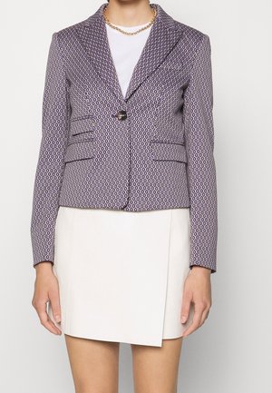 Kobieta ubrana w wzorzysty fioletowy żakiet zapinany na jeden guzik, biały top, białą spódnicę z przednim rozcięciem oraz złoty naszyjnik z łańcucha.