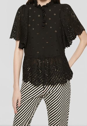 Kvinde iført en sort bluse med hulmønster i blonder med flæset hals og ærmer, kombineret med sort-hvide bukser med diagonale striber.