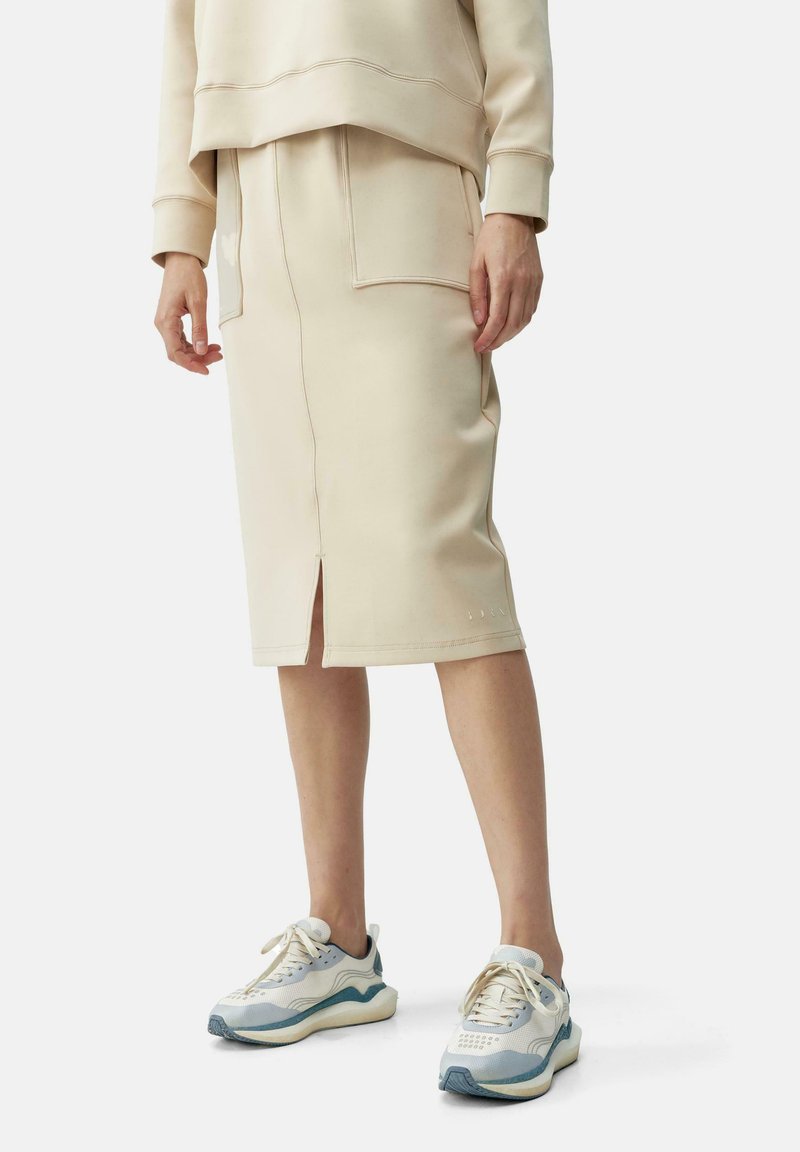 Beige hettegenser og kne-lengde skjørt laget av myk stoff, med frontlommer og en splitt. Kombinert med hvite og lyseblå sneakers.