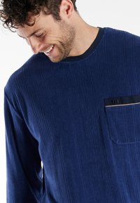 Camisa de manga larga azul marino hecha de un tejido texturizado con rayas verticales y un bolsillo con una línea de acento negra. Cuello redondo.