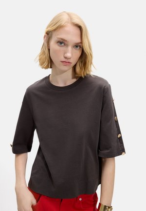 Kobieta o blond włosach do ramion, ubrana w czarny, oversize'owy T-shirt ze złotymi guzikami na rękawach oraz czerwone spodnie, na białym tle.