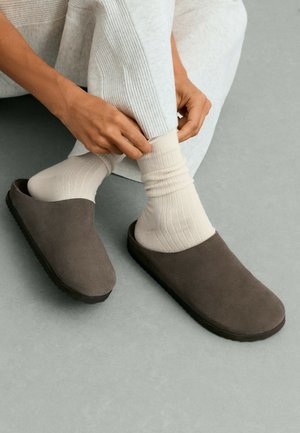 Mains ajustant des chaussettes côtelées de couleur claire portées avec des chaussons gris sans lacets sur un fond de sol gris.
