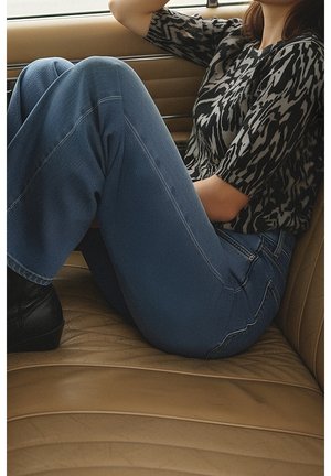 Personne assise de côté sur un siège de voiture en cuir beige, portant un jean bleu, des bottes noires et un haut noir et blanc à motifs.