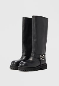 Un par de botas de cuero negro altas con suelas gruesas y hebilla de plata decorativa en las correas del tobillo, mostradas contra un fondo liso.