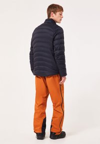 Marineblå polstret jakke med høj krave, prydet med quiltet design. Kombineret med lyse orange bukser og sorte detaljer, vist bagfra.