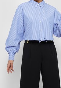 Light blue striped cropped shirt with a button-down collar, balloon sleeves, and a drawstring hem. Paired with black high-waisted trousers.