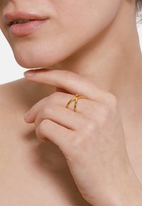Bague en or au design ouvert avec deux bandes entrelacées, reposant sur une main près du menton. Texture lisse et finition brillante.