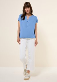Blusa azzurra chiara a maniche corte con colletto a scollatura a V, dettaglio dei bottoni, abbinata a pantaloni bianchi a gamba larga e sneakers beige.