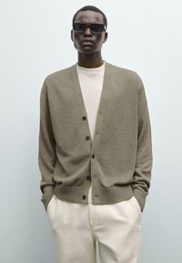 Olivgrüner Cardigan mit V-Ausschnitt, Knopfverschluss und gerippten Bündchen, getragen über einem hellbeigen Hemd; kombiniert mit schwarzen Sonnenbrillen.