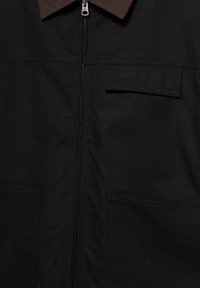 Veste noire avec col marron foncé, fermeture éclair sur le devant, deux poches basses et une poche poitrine à rabat sur le côté droit.