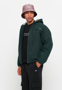 Man wearing a dark green hooded jacket, black HUF shirt, black Champion cargo pants, and a pink bucket hat looking to the side.