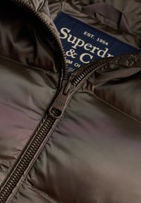 Close-up of partially unzipped black Superdry jacket showing metal zipper and dark blue inner label with white text.
