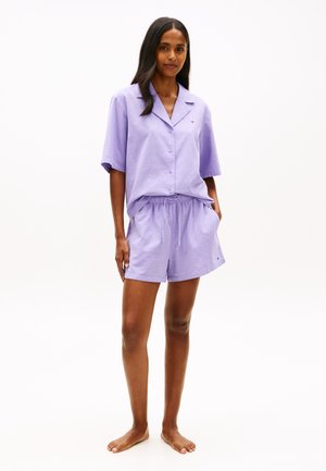 Frau steht barfuß und trägt ein hellviolettes, kurzärmeliges Hemd mit Knöpfen und passende Shorts mit Kordelzug vor weißem Hintergrund.