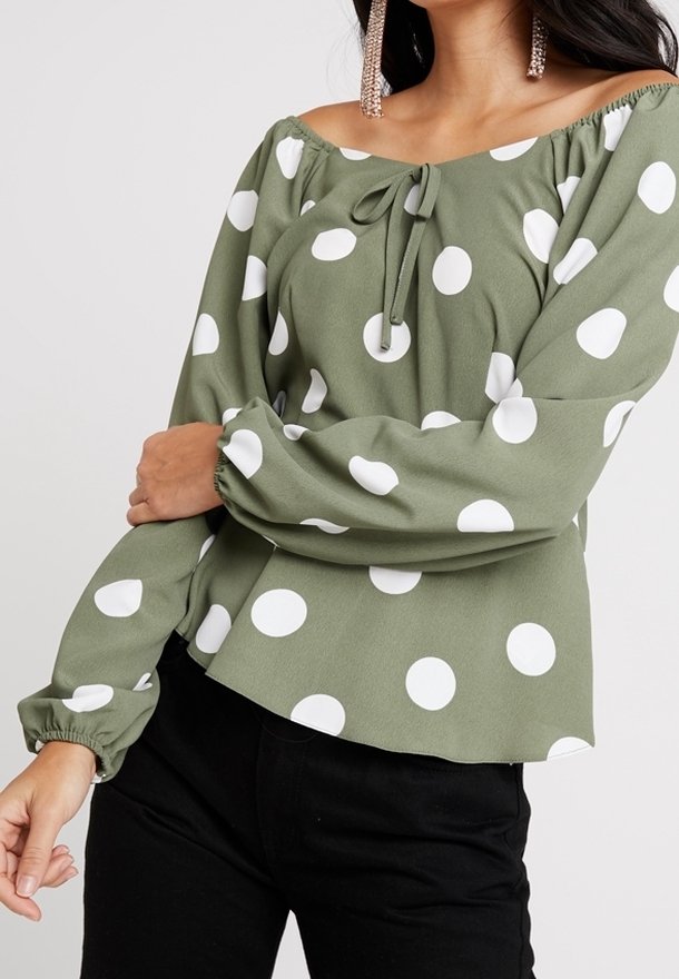 Blusa verde oliva senza spalle con pois bianchi, dotata di scollatura con coulisse e maniche lunghe gatherate, abbinata a pantaloni neri.