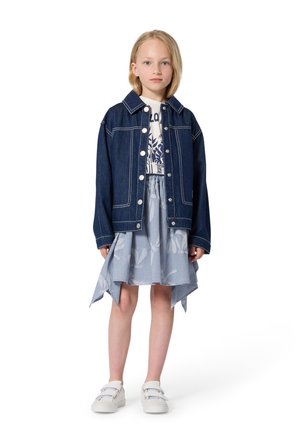 Spijkerjack met witte stiksels over een grafisch T-shirt, gecombineerd met een lichtblauwe asymmetrische rok met bloemenpatroon en witte sneakers.