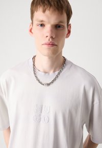 Hvid bomulds T-shirt med broderet "HUGO" logo og en sølv kæde med en vedhæng, der viser en skinnende, refleksiv tekstur.