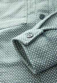 Textured green shirt with a houndstooth pattern, featuring dark buttons and a folded cuff showcasing intricate weave details.