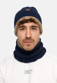 Navy knitted beanie with a brown faux fur cuff and matching ribbed neck gaiter. Both have textured patterns, suitable for cold weather.