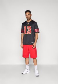 Μαύρη φανέλα NFL με χρυσές και κόκκινες λεπτομέρειες και αριθμό 13, συνδυασμένη με φωτεινά κόκκινα σορτς, λευκά παπούτσια Nike και λευκές κάλτσες.