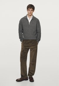 Grå strikket pullover med en halv-zip krave, parret med brune kordfløjlsbukser og mørkebrune sko. Modellen står foran en neutral baggrund.