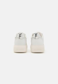 Des baskets blanches avec des accents métalliques sur le talon, une tige en cuir lisse et une semelle en caoutchouc texturée de couleur crème.