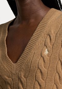 Ruskea neulepaita, jossa on V-pääntie ja ribbireunus; sisältää pienen brodeeratun logon kaula-aukon lähellä; pehmeä rakenne.