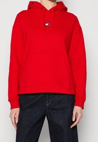 Czerwona bawełniana bluza z kapturem, z przednią kieszenią, sznurkiem do regulacji i naszywką z logo. Posiada ściągacze przy rękawach i u dołu, w zestawieniu z ciemnoniebieskimi dżinsami.