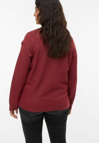 Pull en maille rouge avec un tissu texturé, manches longues, subtil éclat, et ourlet arrondi. Détail supplémentaire : une accent floral sur l'épaule.