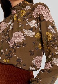 Brun blommig blus med hög smockad krage. Har rosa och gula blommor med gröna blad samt långa ärmar med rynkade muddar.