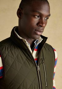 Gep quiltete olivgrüne Weste mit Reißverschluss und seitlicher Tasche, getragen über einem rot-blau gestreiften Hemd mit Kragen. Glatte Textur, schmaler Schnitt.