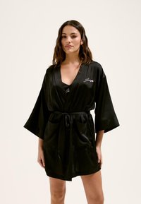 Melna satīna halāta ar saitīti viduklī, platām piedurknēm un kabatām. Iekļauj izšūtu apdari ar vienkāršu burtu uzrakstu priekšpusē.