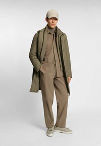 Olivgrüne Daunenjacke über einem beigen Rollkragenpullover, dazu passende hellbraune Hose und cremefarbene Sneakers. Die Mütze ist beige mit Logodetail.