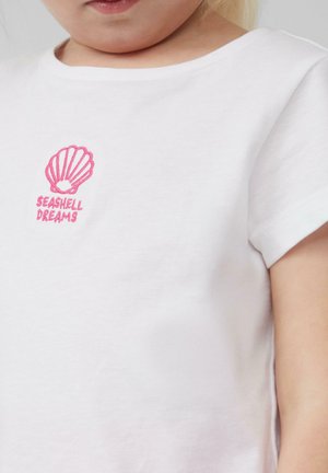 Witte katoenen t-shirt met een roze geborduurd schelpontwerp en de tekst "SEASHELL DREAMS" eronder. Zachte stof, ronde hals, korte mouwen.