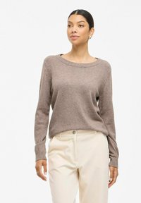 Un pull en tricot marron à manches longues avec un col large et des poignets côtelés, associé à un pantalon côtelé ivoire. Texture douce et coupe décontractée.