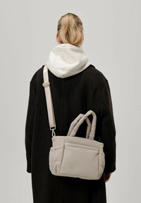 Beige puffer-laukku, jossa on kaksoiskahvat ja irrotettava olkahihna, esitelty tumman takin päällä. Pehmeä, tikattu pinta.