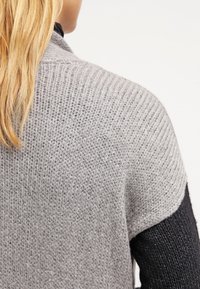 Světlá šedá pletená cardigan s vroubkovanou strukturou, otevřenými rameny a krátkými rukávy, vrstvená přes tmavé tričko s dlouhým rukávem.