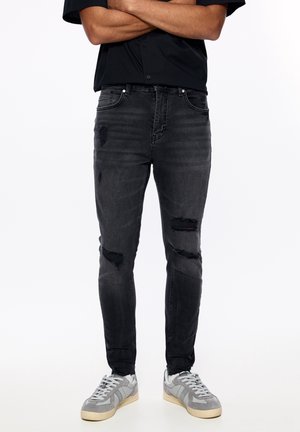 Mężczyzna ubrany w czarne, podarte obcisłe dżinsy, czarną koszulkę z krótkim rękawem i szare trampki, stojący z założonymi rękami.