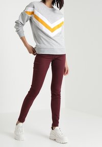 Grijze sweatshirt met gele en witte chevronstrepen, gecombineerd met maroon skinny jeans en witte chunky sneakers. Eenvoudige, casual outfit.