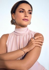 Blusa senza maniche di un delicato rosa pallido con colletto alto, orecchini a cerchio argentati e un semplice braccialetto metallico, abbinati a toni di pelle naturali.