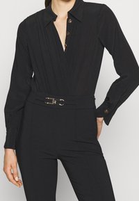 Schwarzer Jumpsuit mit kragenförmigem Ausschnitt, Knopfverschluss, gefältelter Vorderseite und einem metallischen Gürtelakzent in der Taille. Glatte Stofftextur.