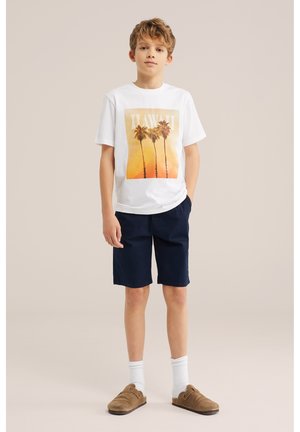 Garçon portant un t-shirt blanc à motif de palmier avec le texte « HAWAII », un short bleu marine, des chaussettes blanches et des chaussures marron sans lacets, debout devant un fond uni.